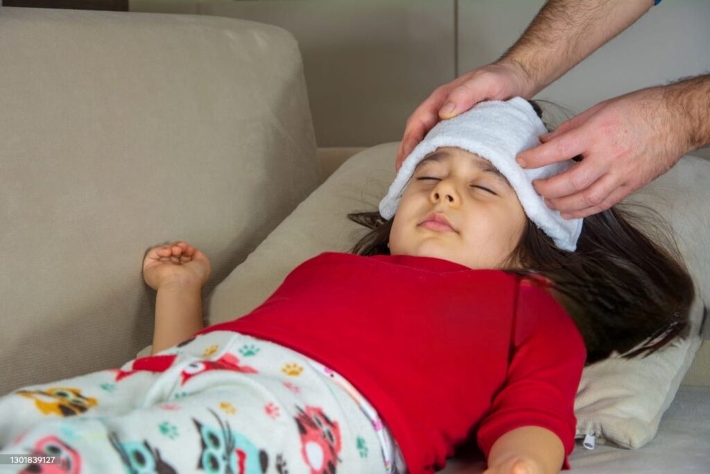 “child lying on sofa with a cold compress on the forehead — fever in children symptoms”